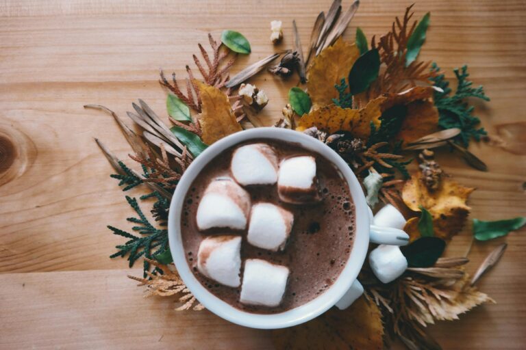 Warm cup of hot chocolate with marshmallows on a wooden table decorated with autumn leaves.